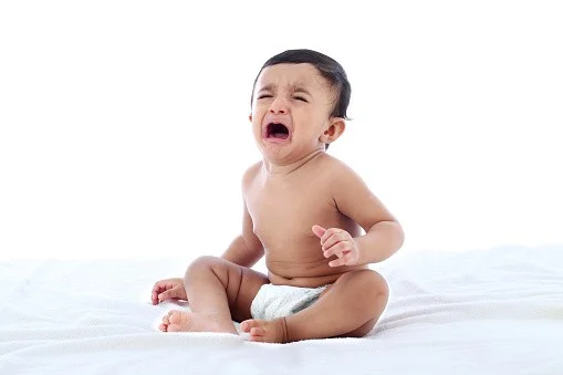 Crying baby sitting on a white surface, representing infant discomfort and the need for chiropractic evaluation.
